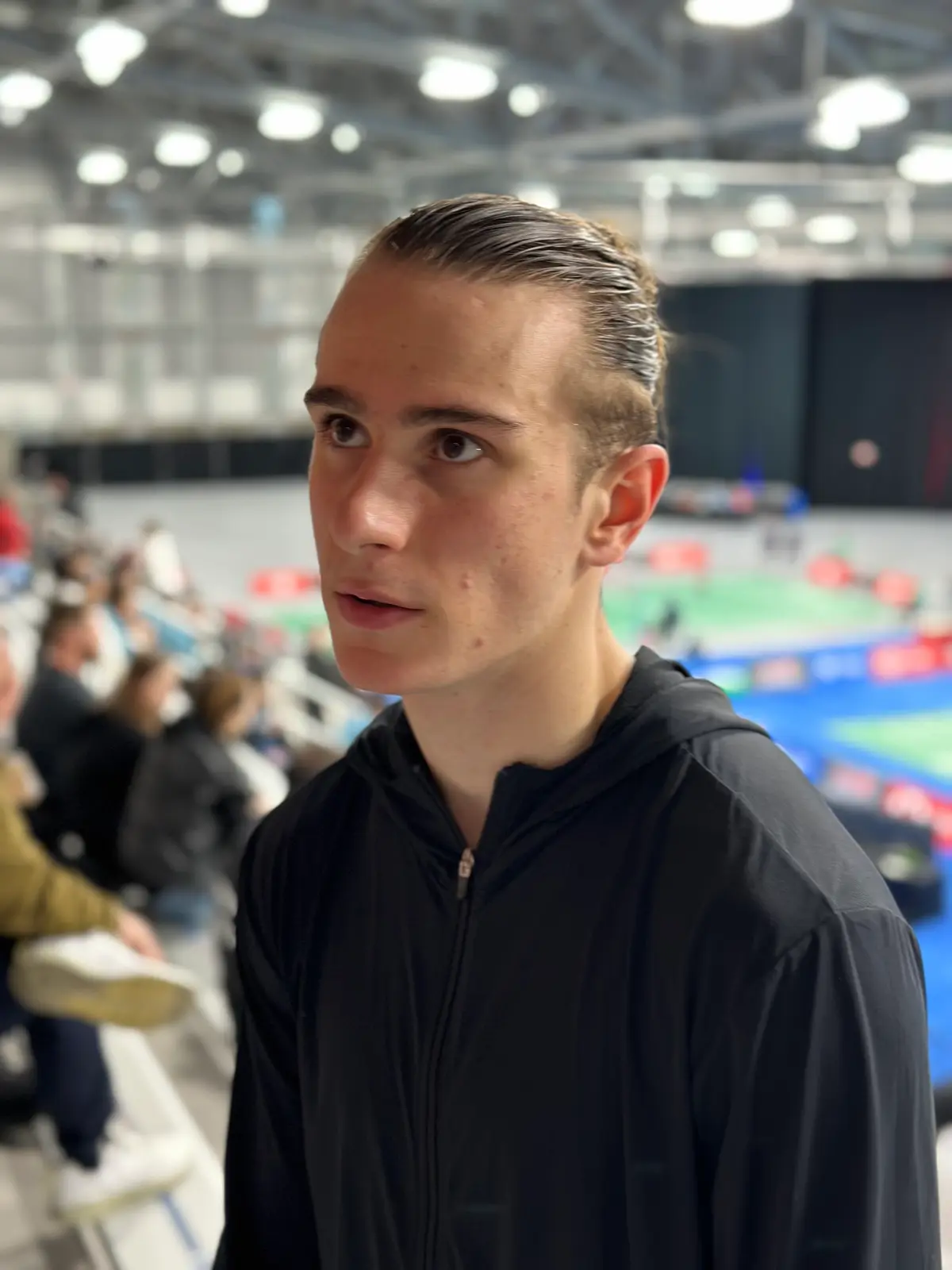 Portrait d'Alex Lanier, cheveux plaqués en arrière et veste noire zippée à capuche, dans les gradins d'une salle de badminton dont les terrains verts et bleus sont flous à l'arrière-plan.